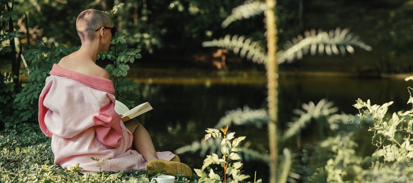 A woman with short hair and bald spots sitting by the lake and reading a book